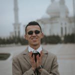 A man in sunglasses and suit smiling in front of a grand mosque with minarets.